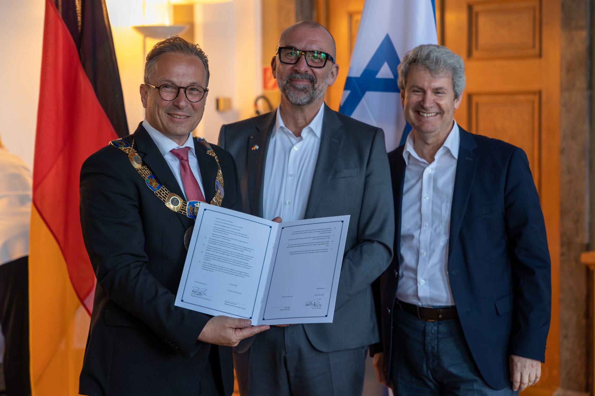 Bürgermeister Reiner Breuer, Bert Römgens und Dr. Oded Horowitz stehen von den Flaggen Deutschlands und Israels. Bürgermeister Breuer hält den unterschriebenen Partnerschaftsvertrag hoch.