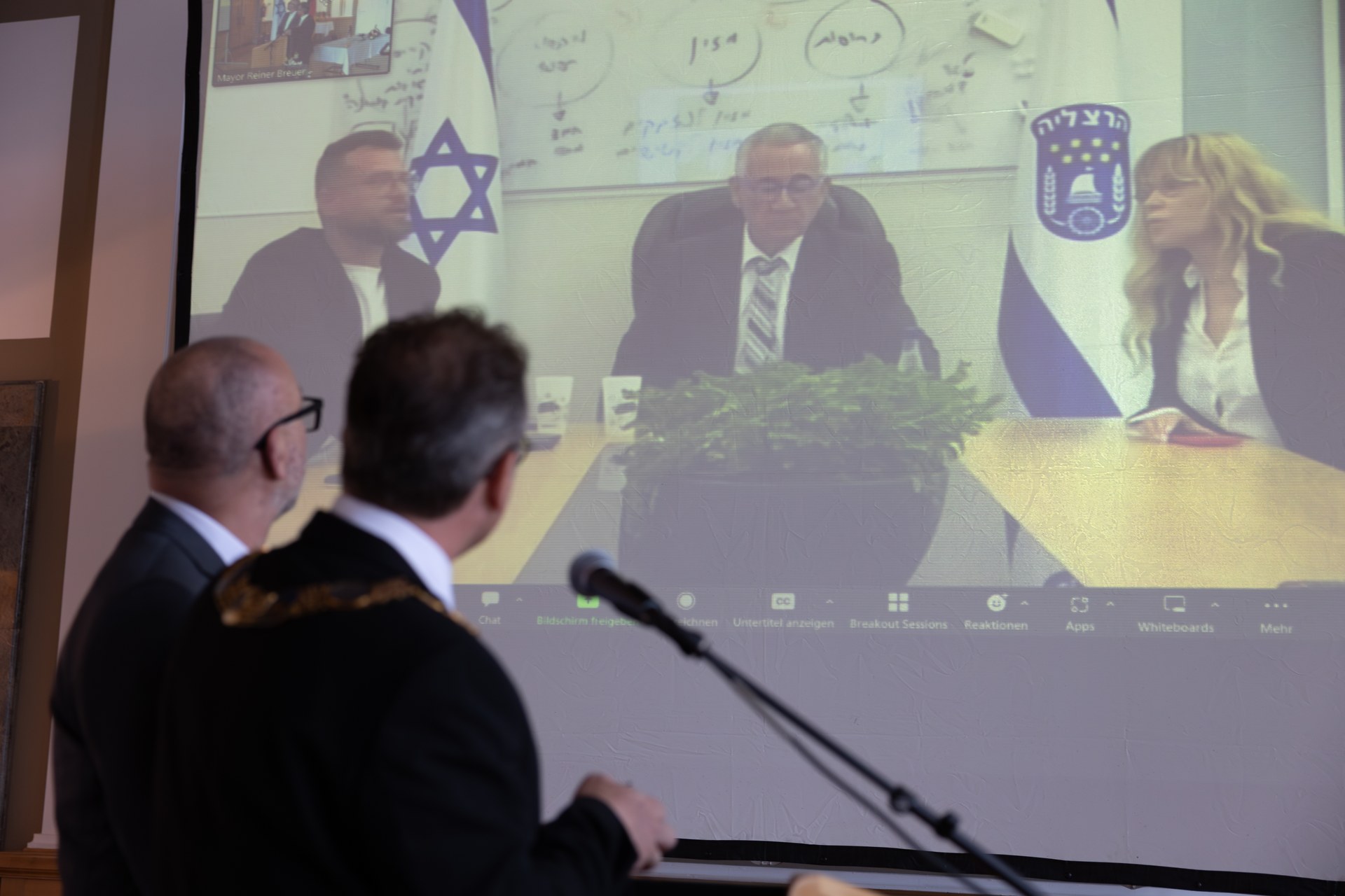Bert Römgens und Bürgermeister Reiner Breuer stehen von einer Leinwand auf welcher der Bürgermeister aus Herzliya Moshe Fadlon sowie die stellvertretende Bürgermeisterin Ofra Bell und der Stadtdirektor Ehud Lazar zu sehen sind.