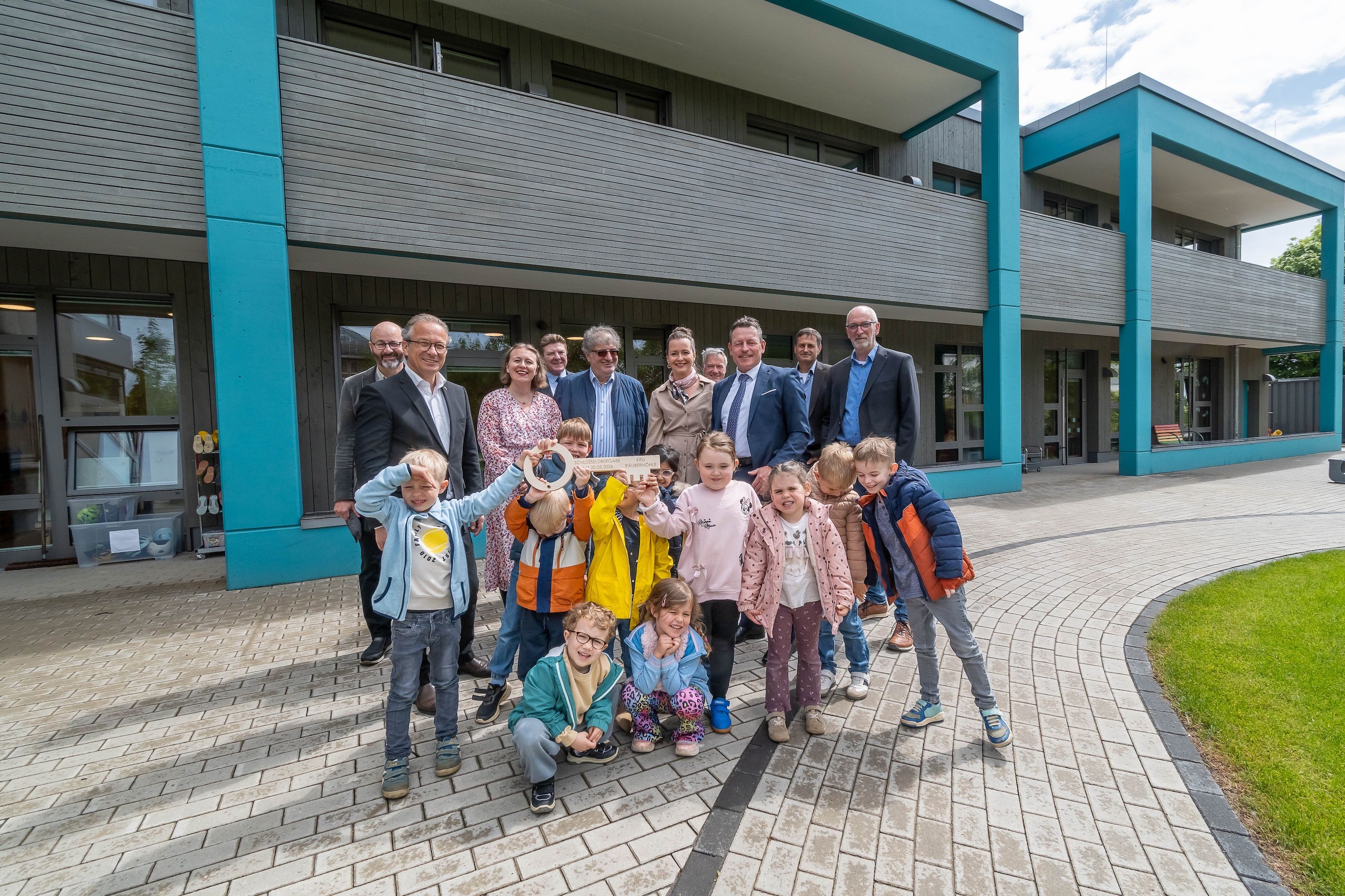 Zu sehen ist eine Gruppe lächelnder Erwachsener, vor ihnen stehen ein paar Kinder im Kita-Alter, die einen großen Schlüssel in der Hand halten. Im Hintergrund ist ein modernes, zweigeschossiges Gebäude zu sehen