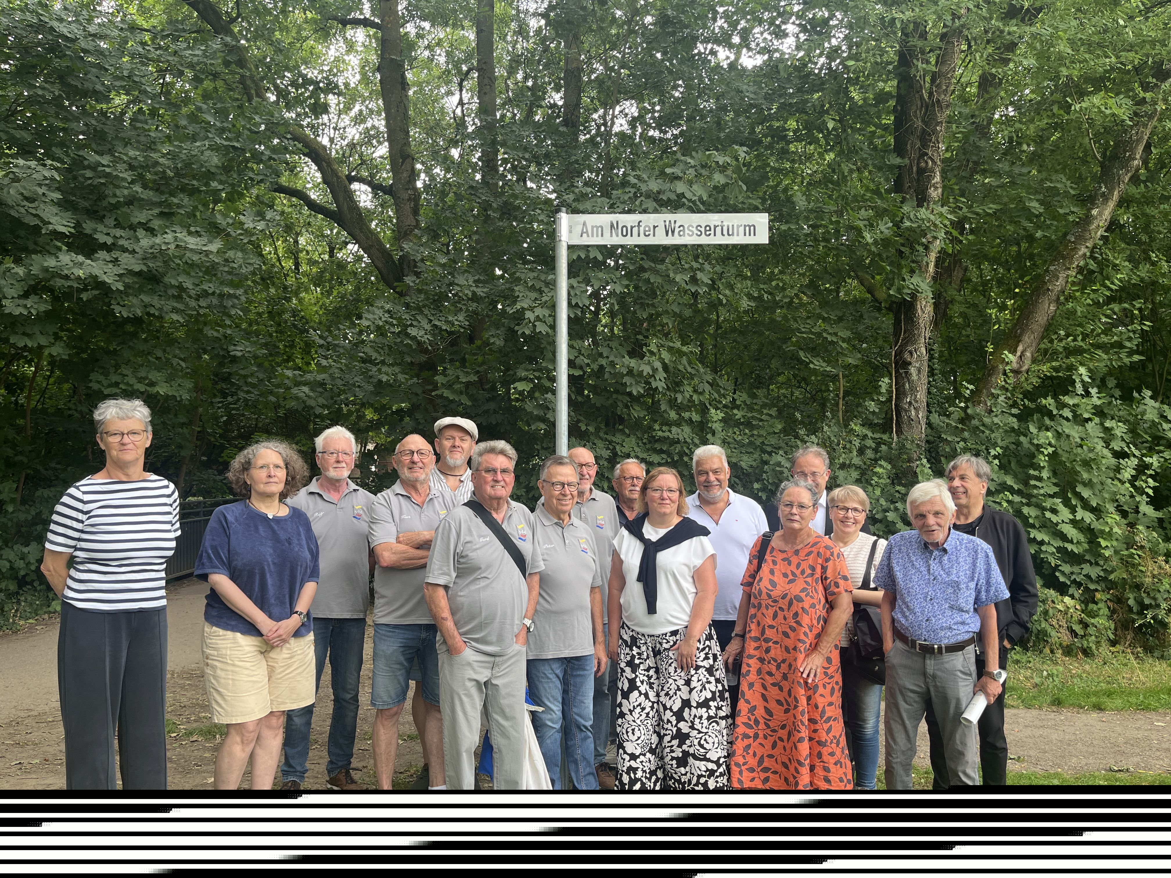 Eine Gruppe Erwachsener steht lächelnd auf einer Wiese, hinter ihnen ein Straßenschild