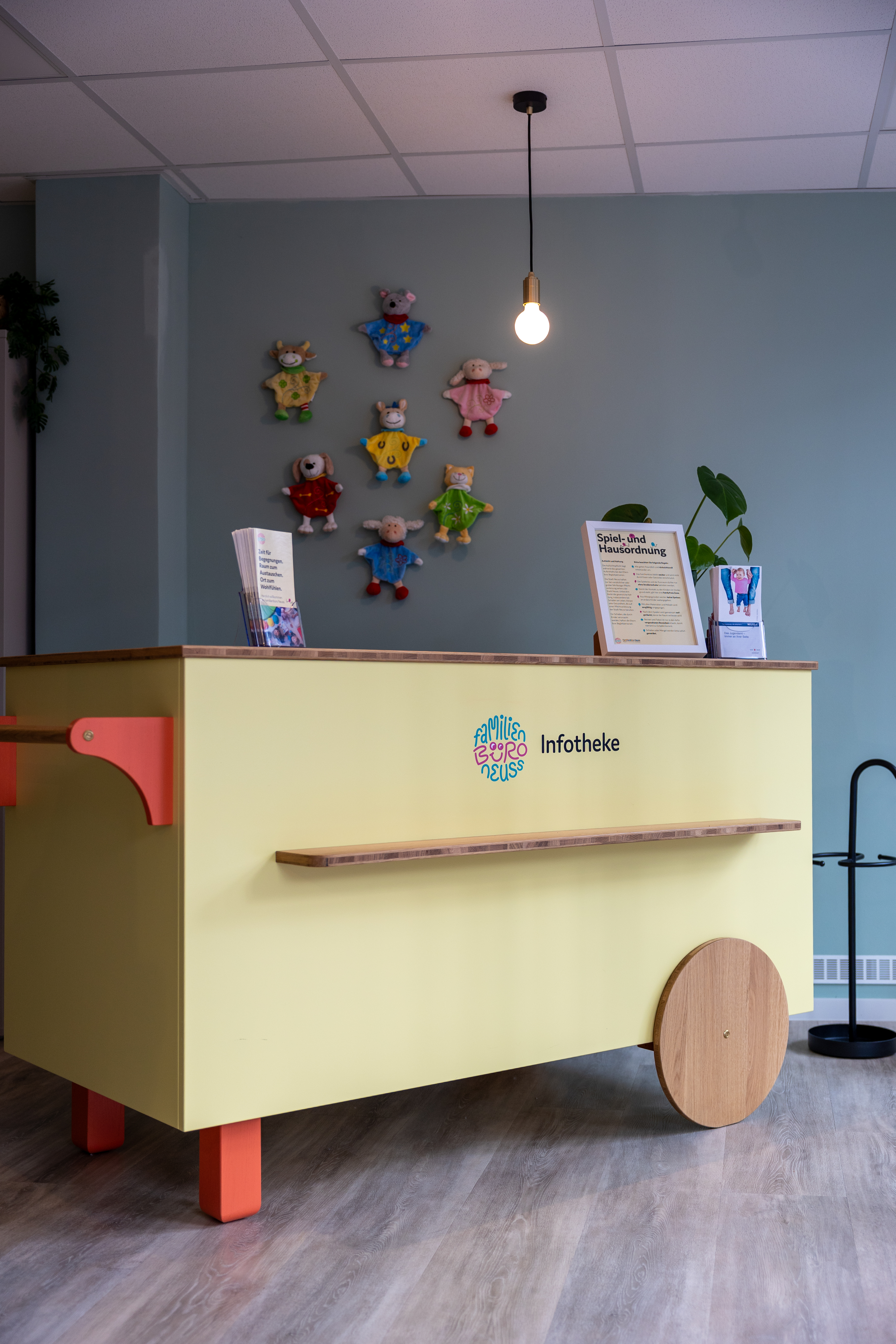 Inneaufnahme, im Zentrum des Bildes ist eine hellgelbe Infotheke, darauf stehen Flyer und eine Pflanze. Im Hintergrund ist Spielzeug zu sehen.
