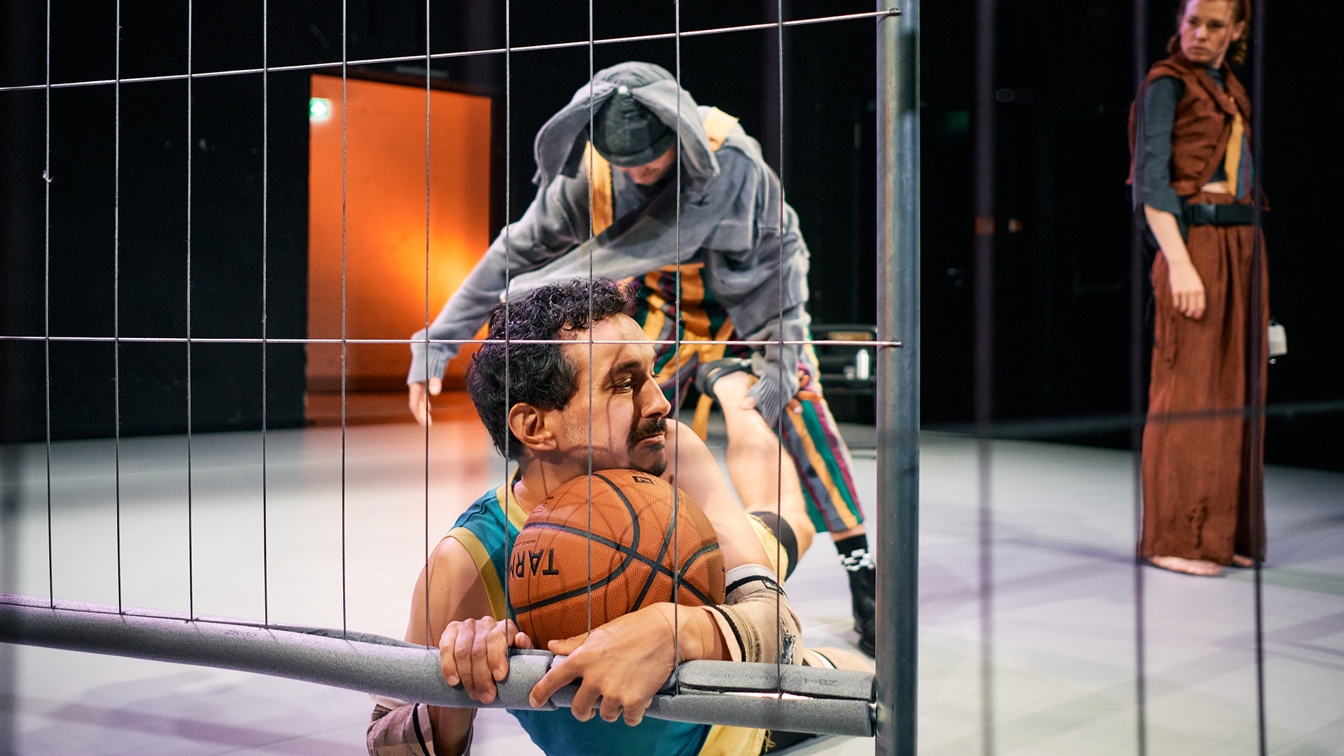 Ein Theaterstück mit drei Schauspielern, einer in einem Basketballtrikot, hinter einem Gitter.