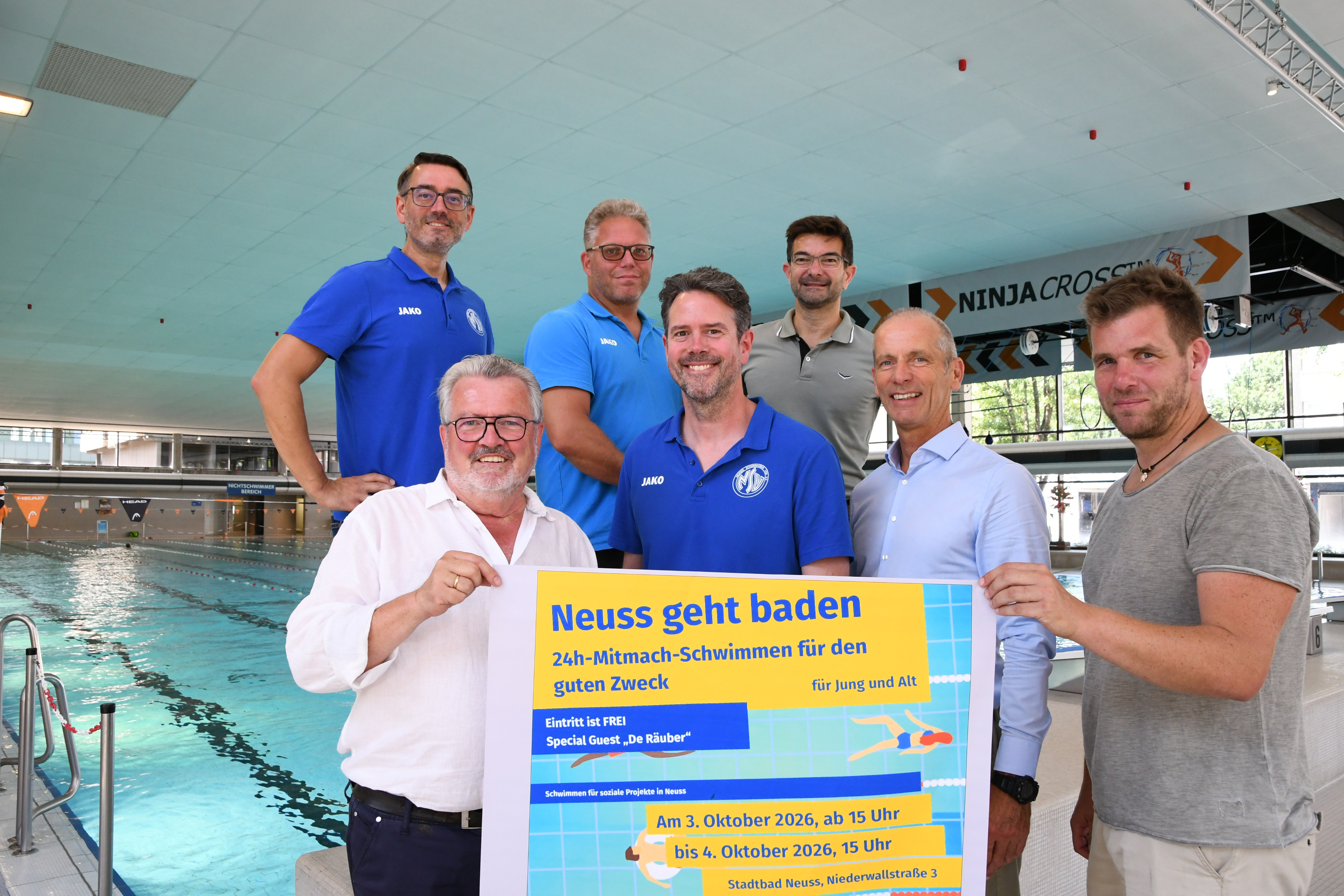 Eine Gruppe von Menschen steht im Schwimmbad, schaut in die Kamera und präsentiert ein Plakat zum 24-Stunden-Schwimmen.