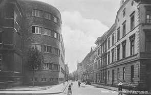 Schwarz-Weiß-Aufnahme des Anbaus mit Blick auf die Kanalstraße, auf der mehrere Menschen zu Fuß oder mit dem Fahhrad unterwegs sind. Ein einziges geparktes Auto ist zu sehen.