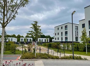 Straßenansicht eines modernen Wohnkomplexes mit einem Spielplatzbereich. Auf dem Spielplatz sind unter anderem eine Schaukel und eine Rutsche erkennbar.