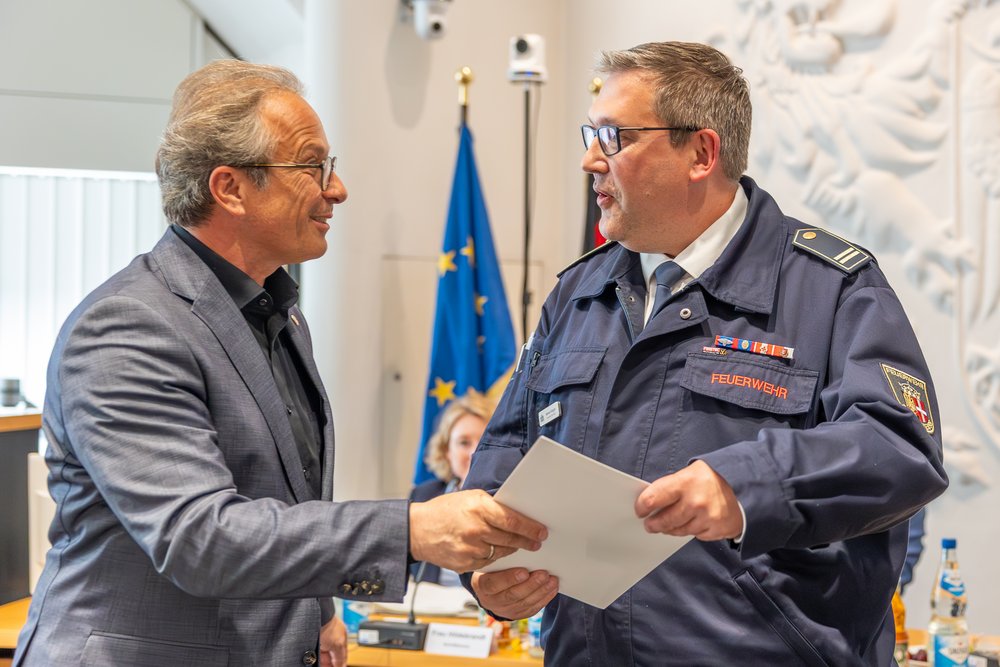 Innenraum. Ein Mann in Anzug übergibt einem Mann in Feuerwehruniform eine Urkunde, sie geben sich lächelnd die Hand.
