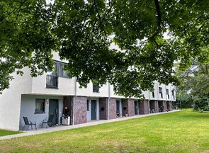 Rückseite eines Reihenhauses mit mehreren Hintereingängen, jeweils mit einem kleinen Terrassenbereich und umgeben von einer Grünfläche.