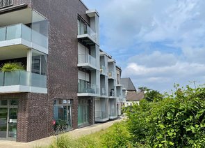 Rückseite zweier moderner Mehrfamilienhäuser mit Balkonen, eines mit dunklen Ziegeln versehen, das andere hell verputzt. Daneben ist ein Grünbereich mit Rasen und dichten Büschen zu erkennen.