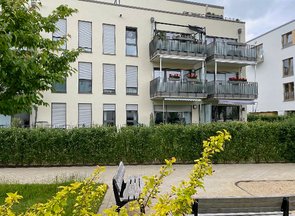 Rückansicht einer modernen Wohnanlage mit Balkonen einer grünen Hecke davor. Im Vordergrund sind ein Gehweg und zwei Sitzbänke zu sehen.