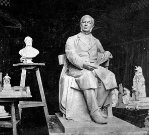 Eine erkennbar kleinere Version des Denkmals steht in einem dunklen Raum. Im Hintergrund ist eine Miniatur des Denkmals, sowie eine Büste Theodor Schwanns zu erkennen.