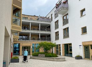 Innenhofartiger Eingangsbereich eines modernen Wohngebäudes. Davor befindet sich eine ältere Person mit einem Rollator. An der hellen Fassade des Gebäudes befinden sich Balkons und Erker. Vor dem Eingang ist eine Grüninsel mit Sitzflächen erkennbar.