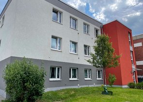 Vorderfront eines zweigeschossigen grauen Wohngebäudes mit rot abgesetztem Eingangsbereich und einer Grünfläche im Vordergrund.