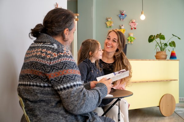 Eine Frau sitzt mit ihrer Tochter auf dem Schoß an einem kleinen Tisch und lässt sich von einem Mann beraten.