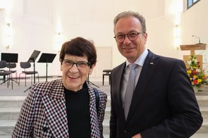 Frau Süssmuth und Herr Breuer lächeln in die Kamera – im Hintergrund ist der sonnenlichtdurchflutete Konzertsaal des Zeughauses zu sehen.