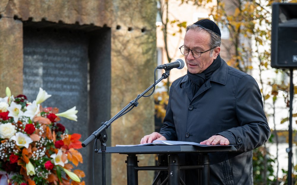Bürgermeister Reiner Breuer spricht an einem Podium -- im Hintergrund sieht man das mit Blumen gesäumte Mahnmal an der Promenadenstraße.