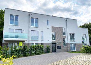 Frontansicht eines modernen Mehrfamilienwohngebäudes mit hellgraue verputzten Wänden, geklinkerten Akzenten und einem Balkon.