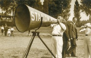 Ein Mann steht hinter einem kegelförmigen Sprachrohr und macht scheinbar eine Durchsage. Das Sprachrohr ist fast so groß, wie der Stadionsprecher und wurde folglich auf ein Stativ montiert.