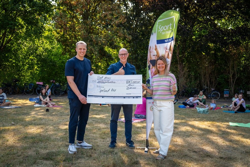 Drei Personen mit einem Geldcheck vor einer "Sport im Park"-Fahne. 