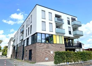 Blick von der Straße auf ein modernes Wohngebäude mit Balkonen, teils geklinkerter Fassade und gelben Farbakzenten an den Fenstern.