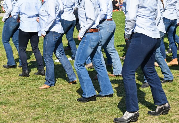 Eine Gruppe von Line-Dancern tanzen auf einer Wiese. Es sind jeweils fast ausschließlich die Beine zu sehen – natürlich stilsicher in Blue Jeans und Cowboystiefeln.