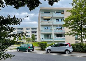 Blick von der Straße auf win Wohnhaus mit Balkonen. Davor befinden sich geparkte Autos, dahinter weitere Wohngebäude.