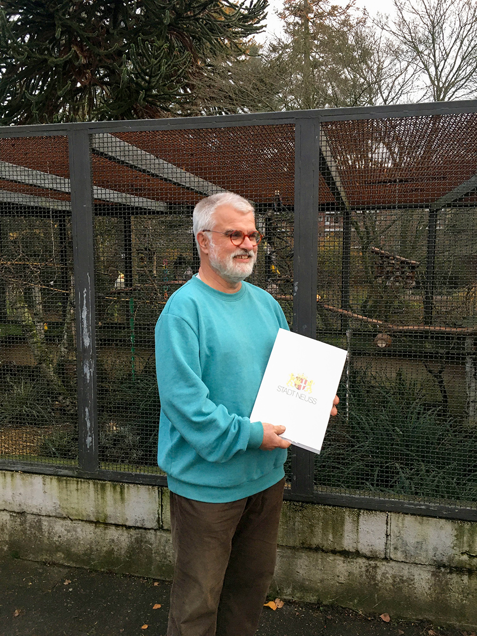 Gärtner Reinhard Gremmer vor der Vogelvoliere. Er lächelt und hält eine Mappe mit der Urkunde für seine 40 Dienstjahre in den Händen.